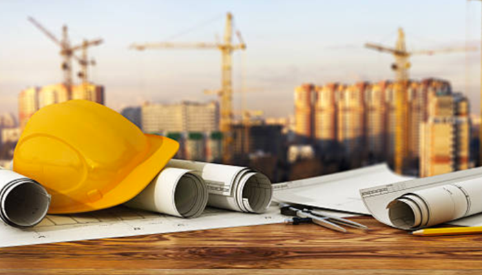 a hard hat and set of paper plans lying on a desk with construction cranes in background